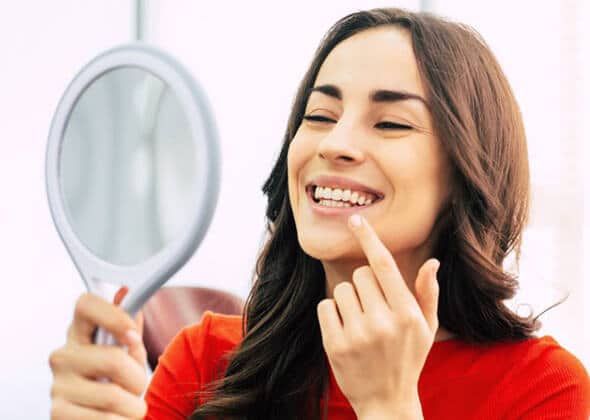 Woman in red shirt smiling at mirror, touching her teeth and face.