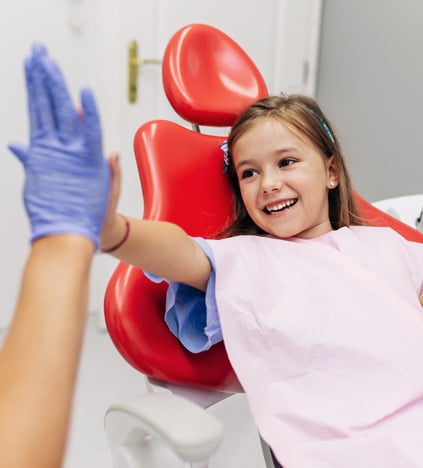 Young girl wearing red boxing gloves playfully punching with adult wearing blue gloves in dental office.