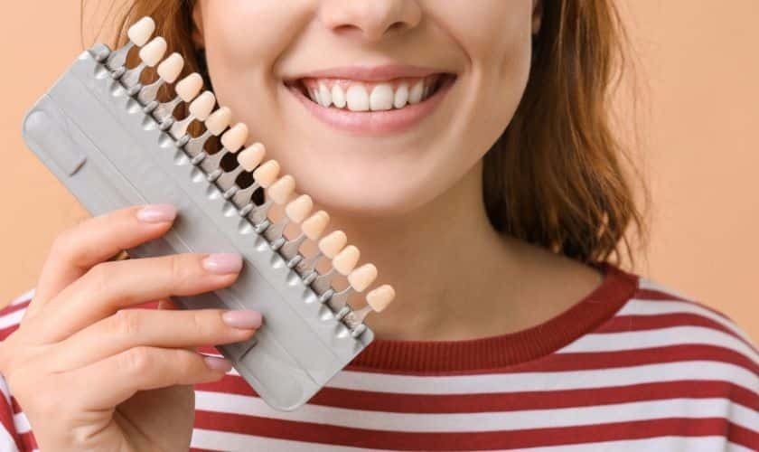 Woman smiling while holding a dental shade guide showing tooth color options for veneers.