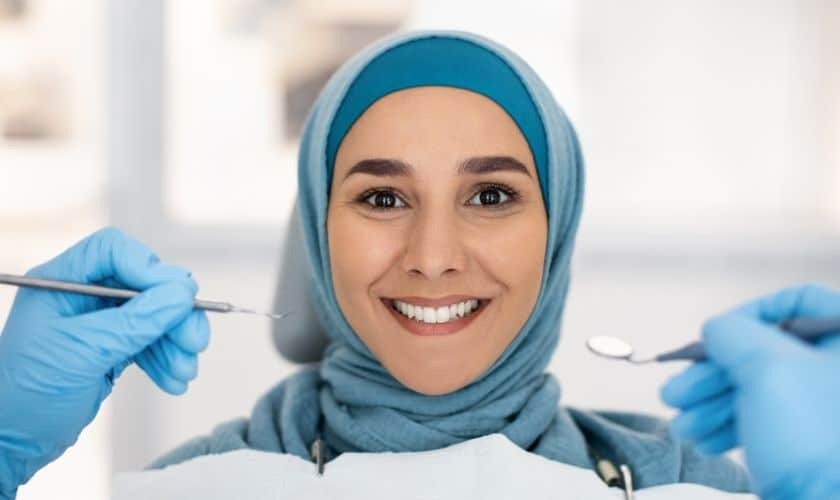 Smiling female dentist in blue hijab and gloves holding dental instruments during cosmetic treatment.